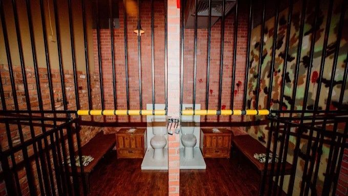 Prison cell interior: two toilets, barred walls, wooden benches, and brick background.