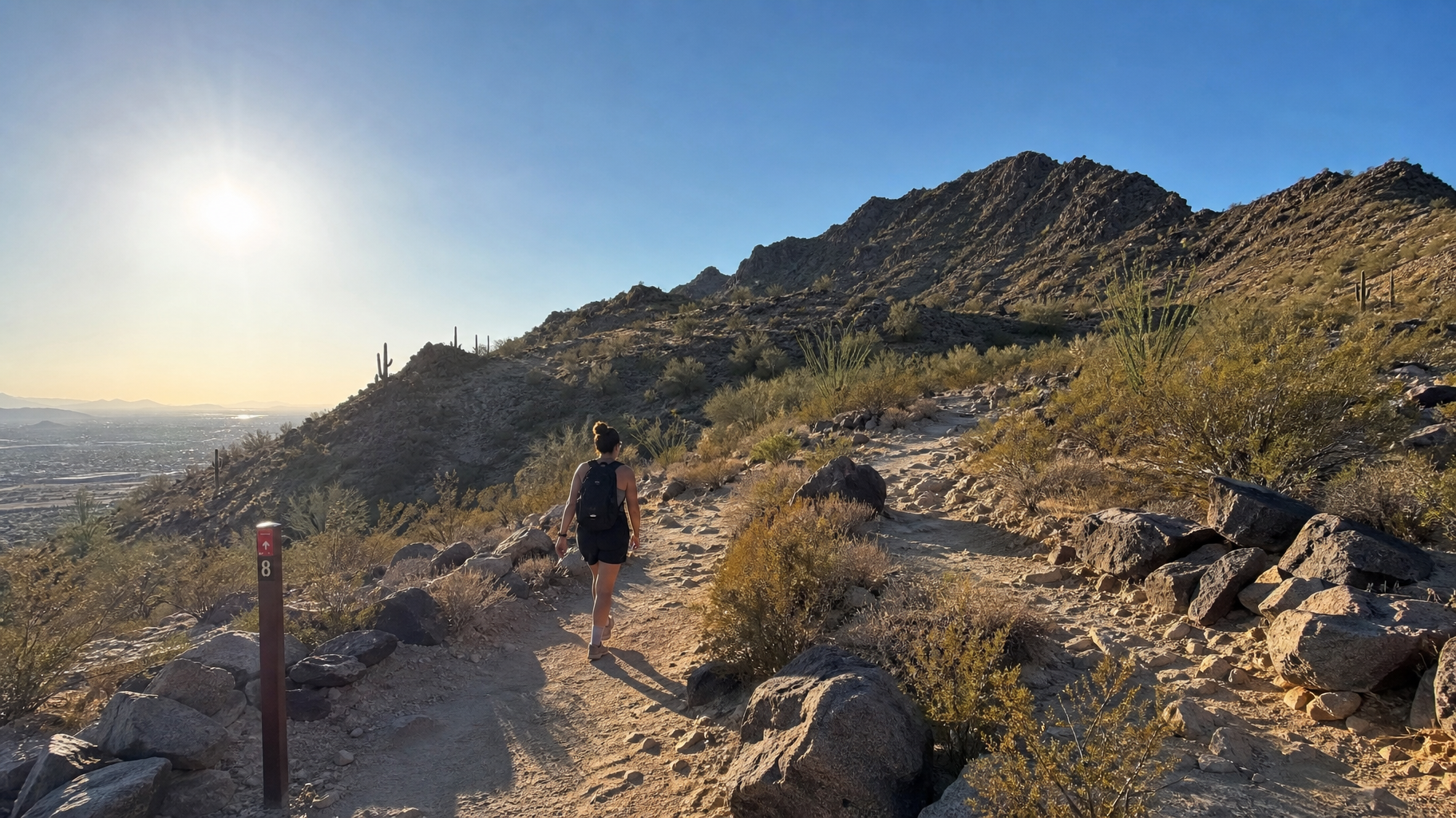 Hiker on a rocky desert trail at sunset with mountains and city lights in the distance