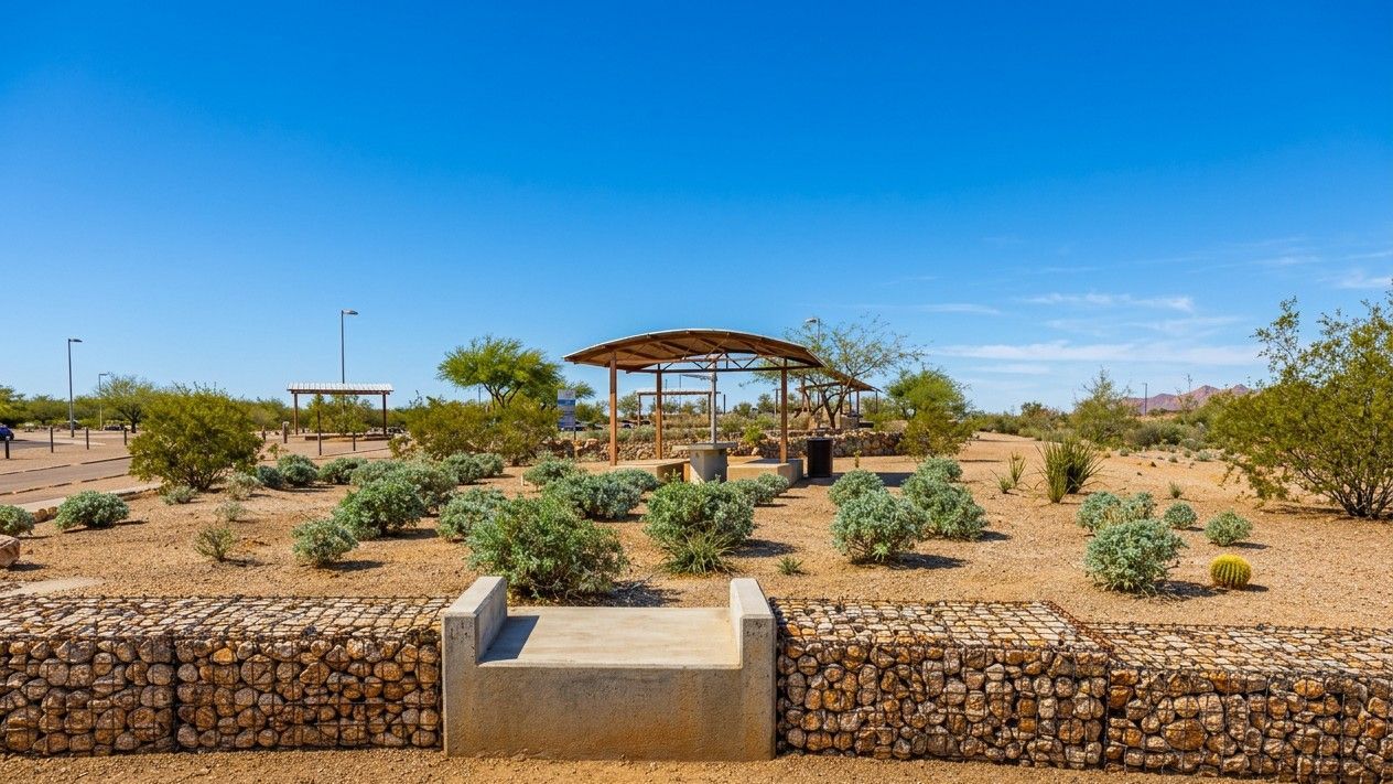 Desert garden with a stone wall, shrubs, and a shaded pavilion under a bright blue sky.