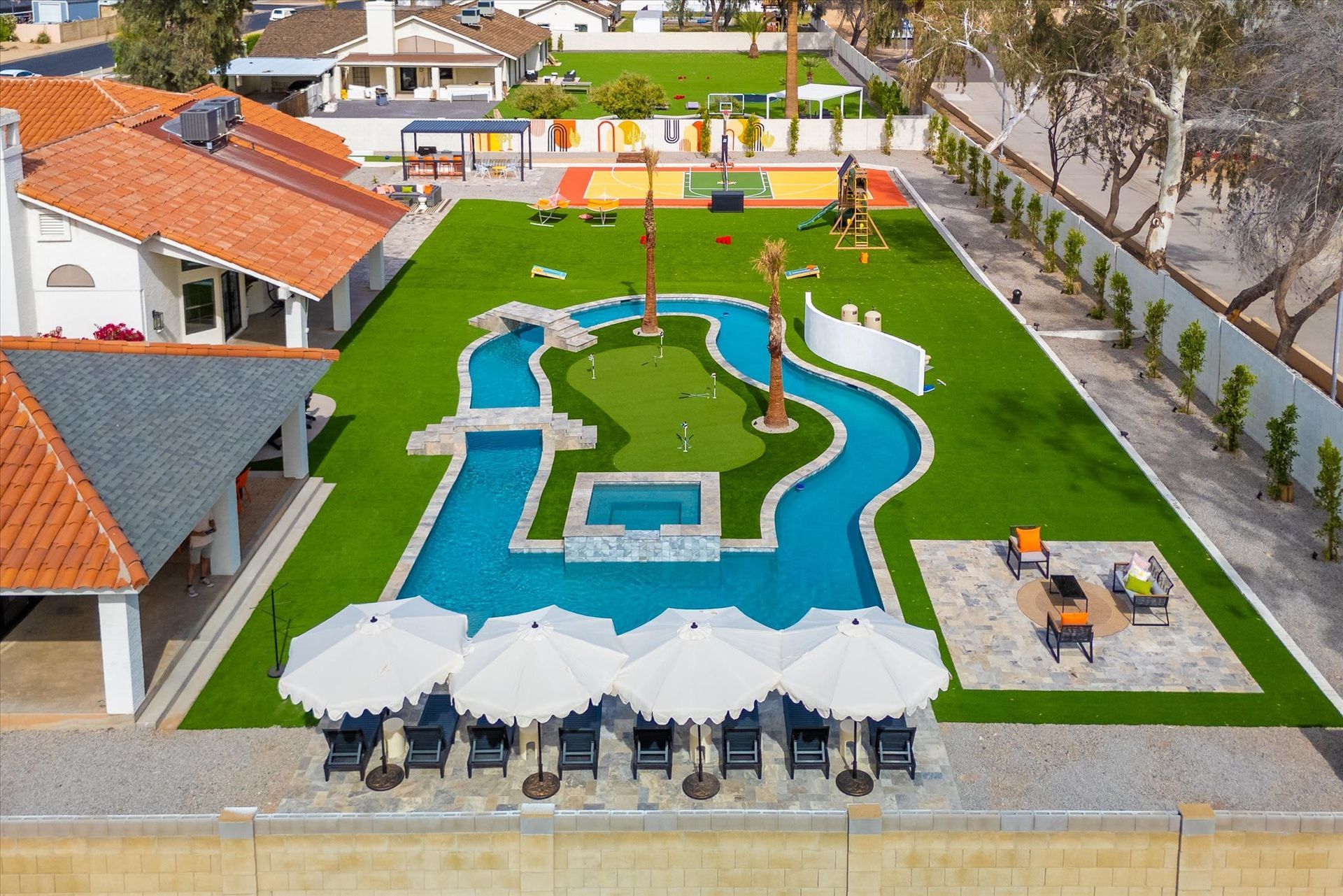 Aerial view of a resort pool with winding blue water, white umbrellas, green lawn, and red-roofed buildings