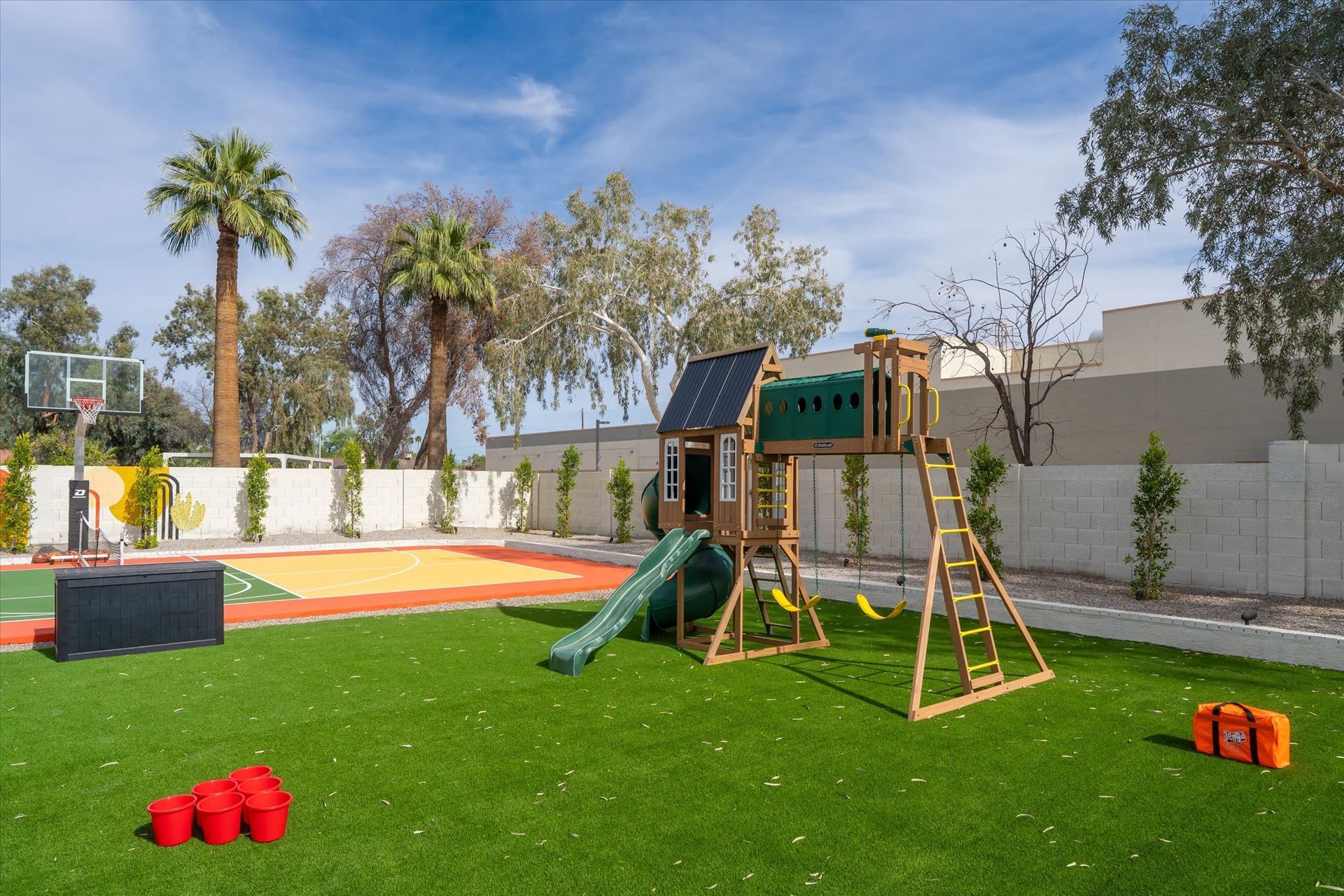 Backyard playground with green turf, slide, swings, palm trees, and a small trampoline under a blue sky