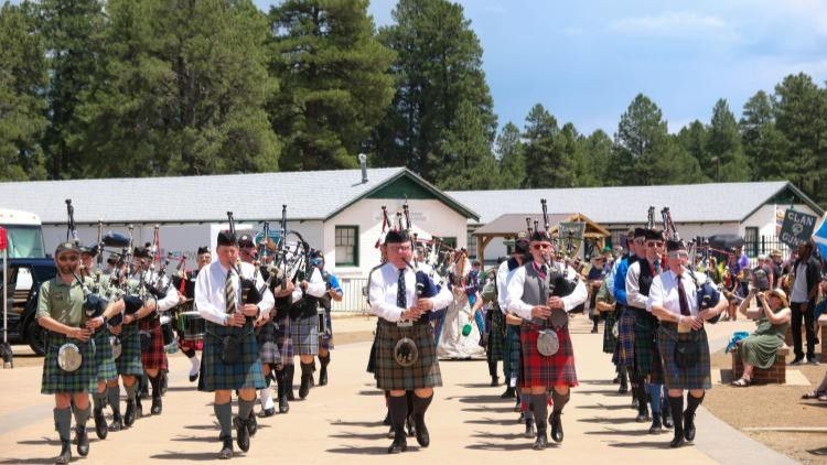A pipe band in kilts and formal uniforms marches down a path in front of a white building surrounded by pine trees.