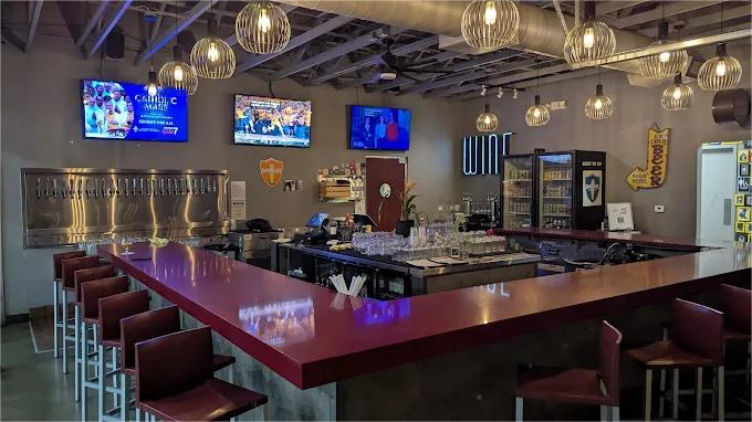 Bar interior with a long red counter, stools, beer taps, TVs, and globe lights.