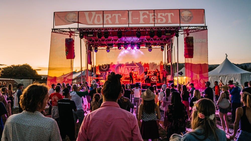 Crowd at outdoor festival stage under sunset sky. People dance, band plays on stage with lights.