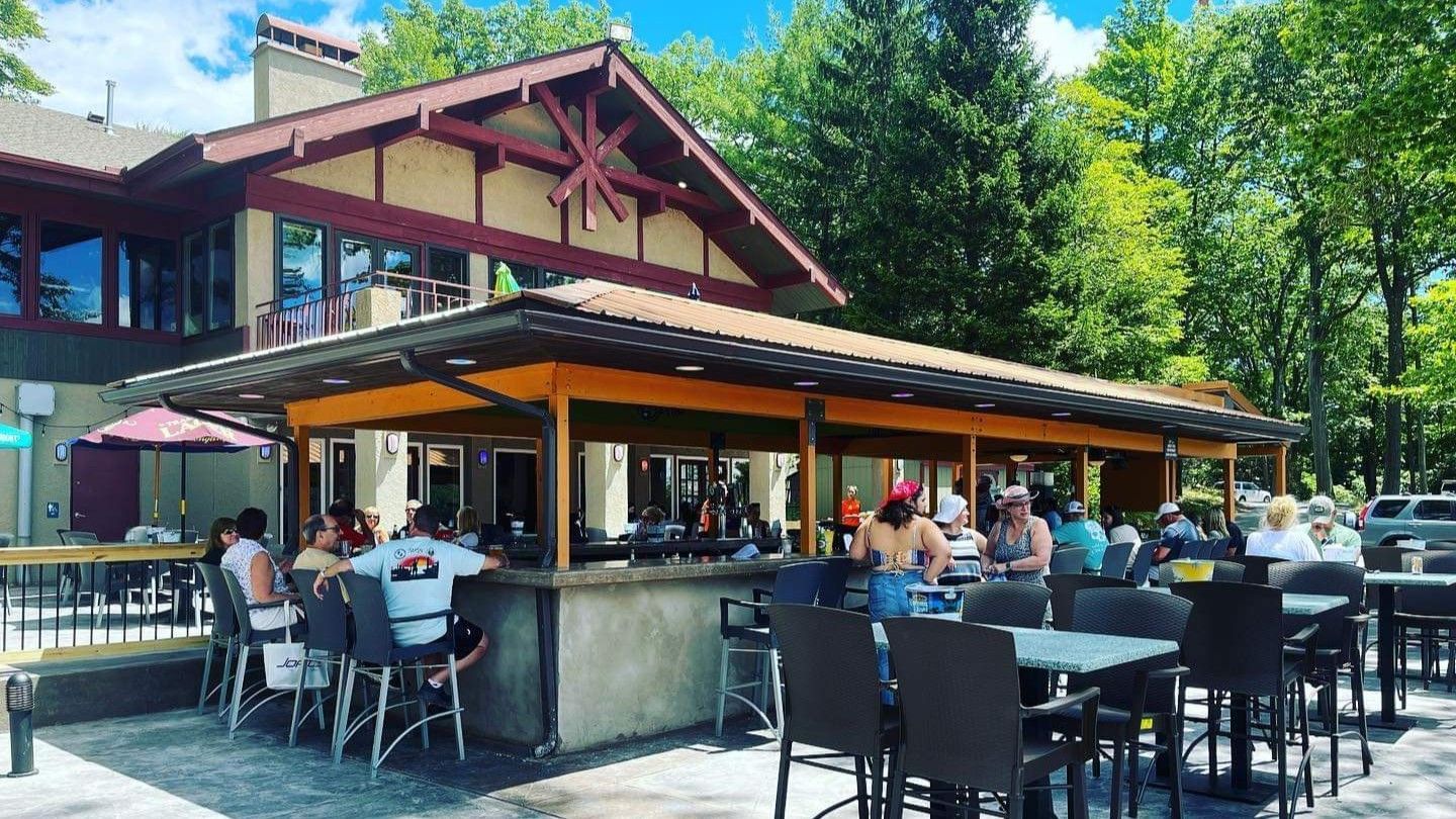 An outdoor bar and restaurant with people seated at tables, a covered bar, and a building with a brown roof in a wooded area.