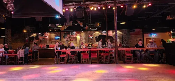 A dimly lit country western bar with people at tables and a dance floor. Red chairs and a wooden bar.