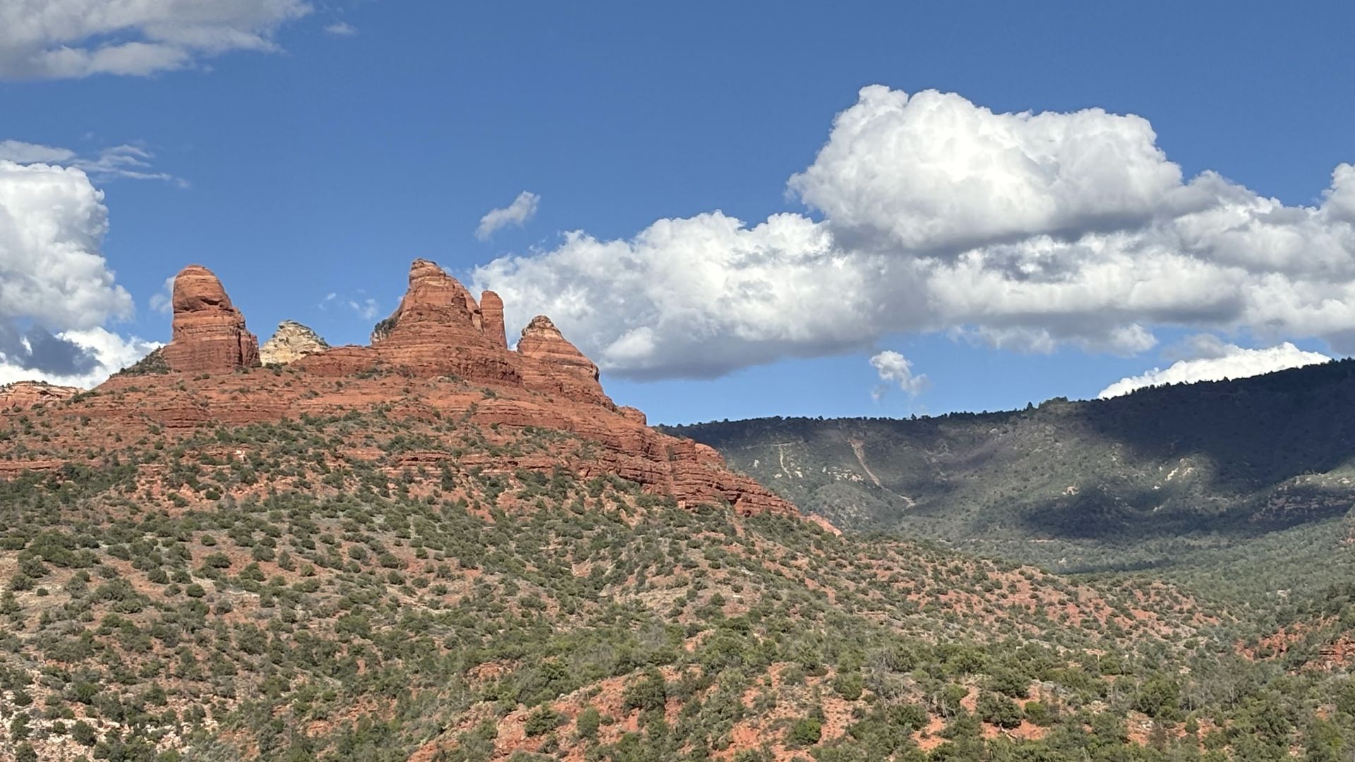 Red rock formations and scrub brush landscape under a blue sky with fluffy white clouds.