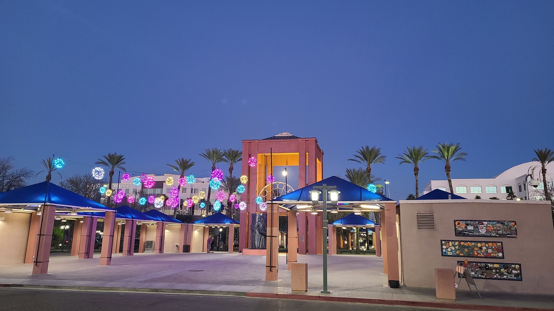 Outdoor plaza at dusk with illuminated structures and palm trees.