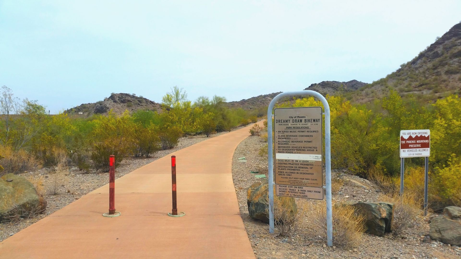 Concrete path through a desert landscape with signs and yellow bushes.
