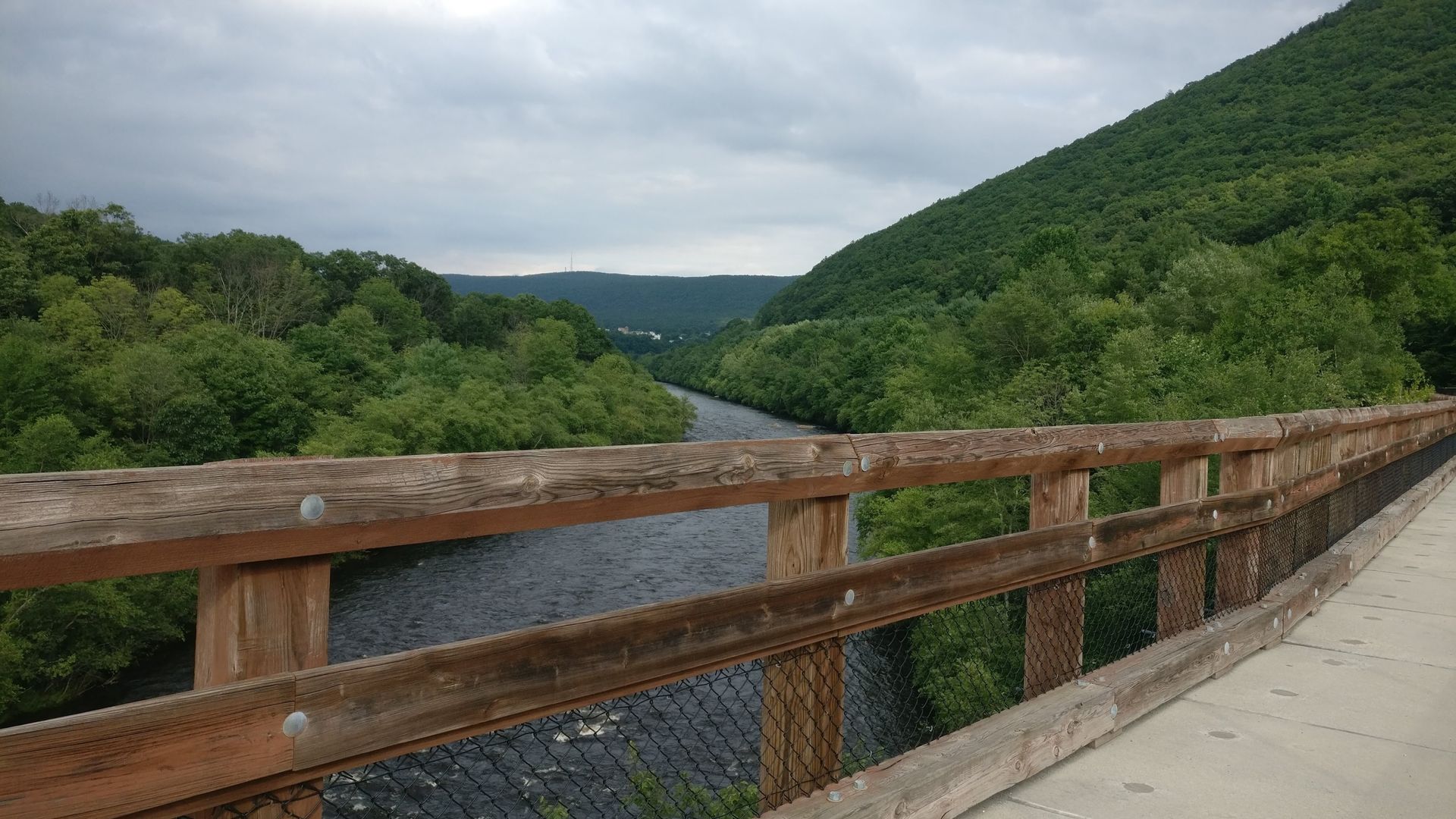 Wooden bridge over river surrounded by green trees and mountain under cloudy sky.