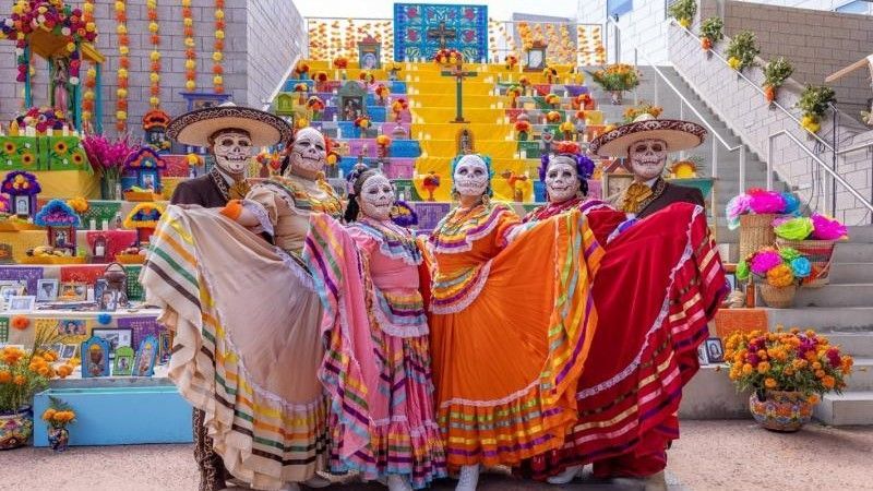 People in Day of the Dead costumes pose in front of a colorful altar with flowers and decorations.