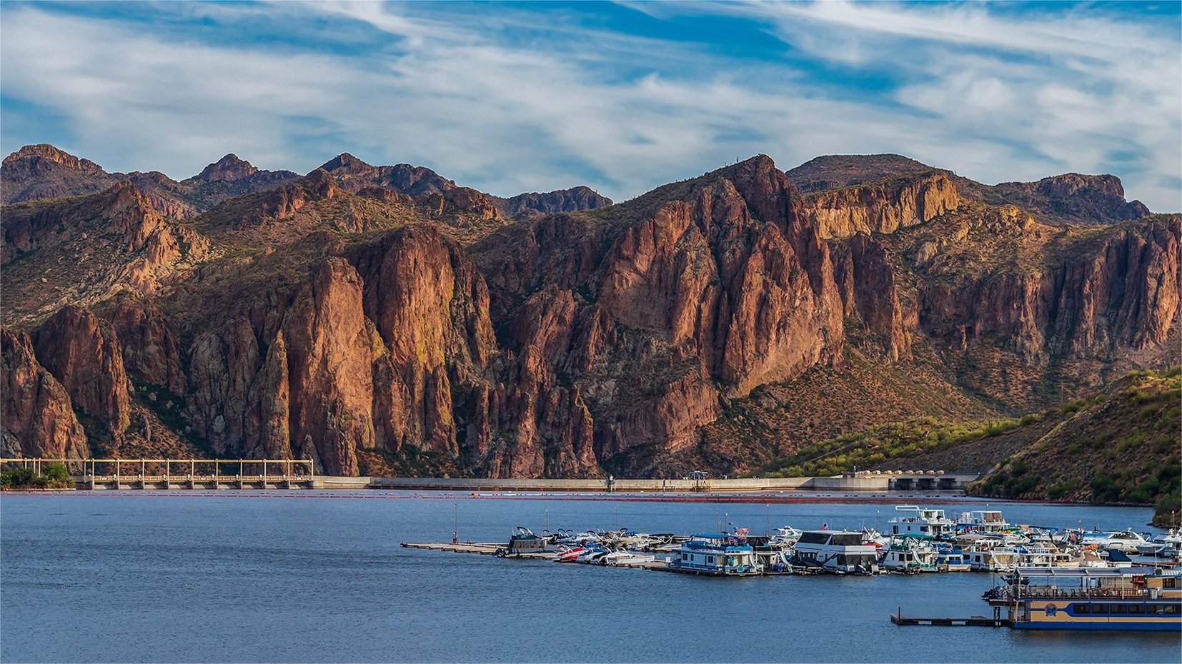 Lake with boats and mountains under a blue sky. A dam is visible in the background.