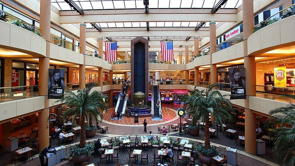 Interior of a multi-level shopping mall with stores, escalators, and a central fountain area. Palm trees and American flags present.