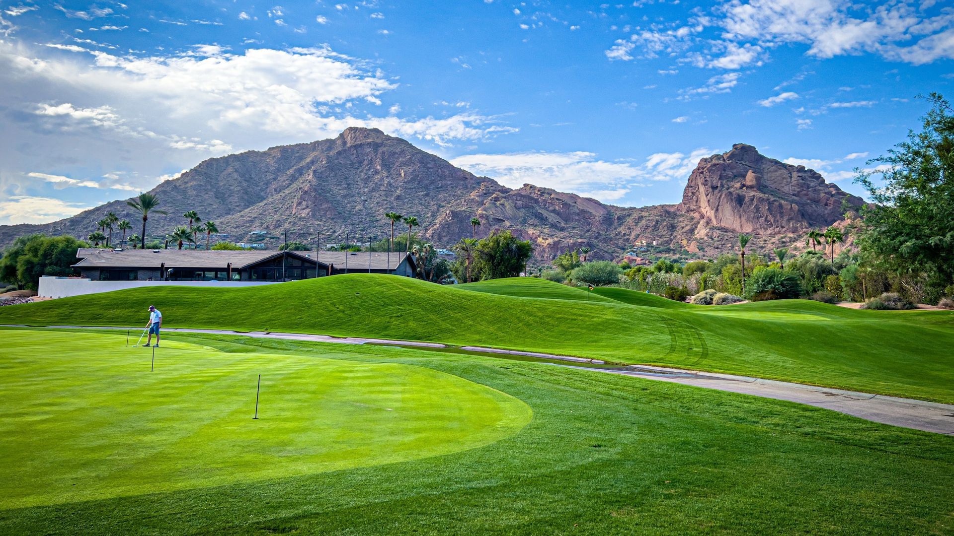 Green golf course with mountains in the background under a blue sky. Person is putting on the green.