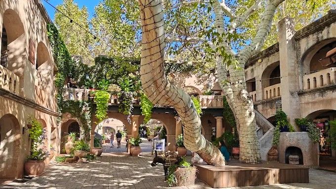 Courtyard with arched walkways, large tree, and people. Buildings are tan, with greenery.