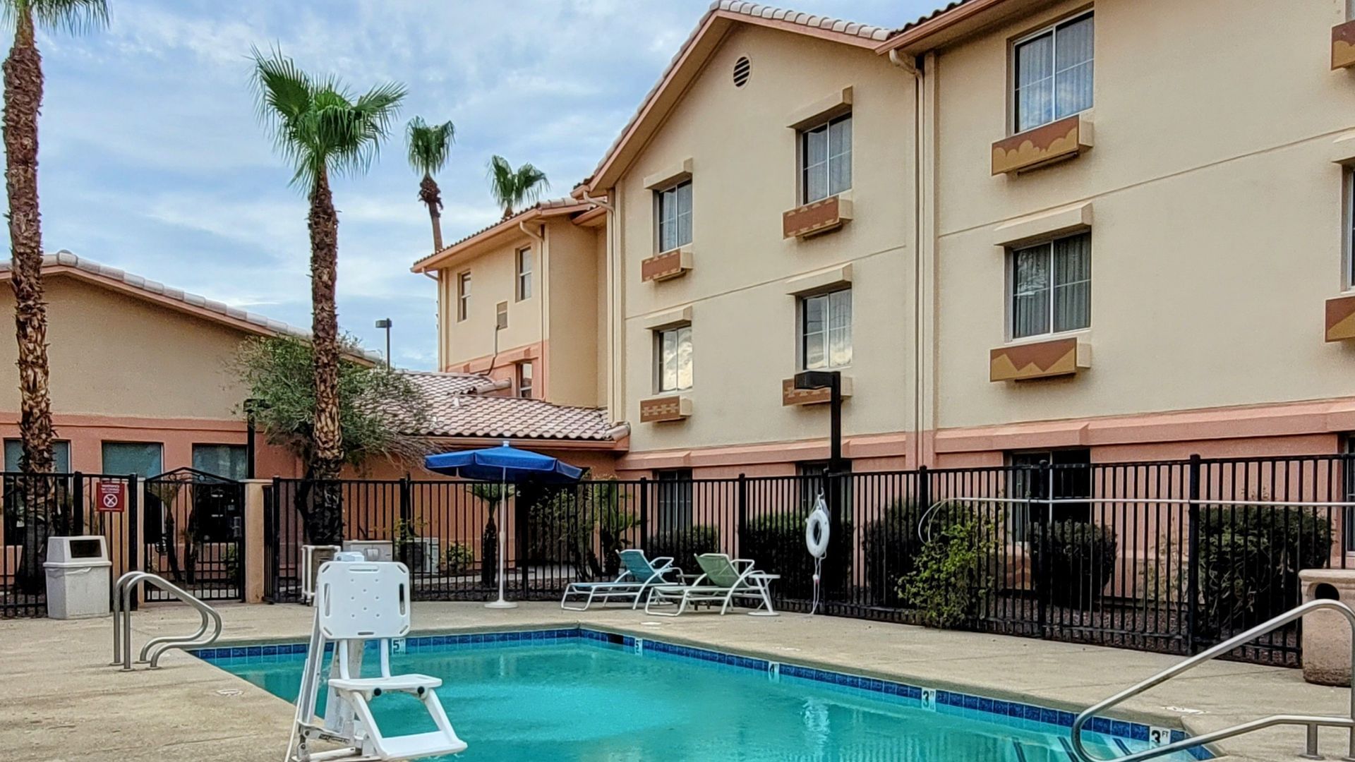 Hotel exterior with pool, palm trees, and overcast sky. Building is tan with red roof accents.