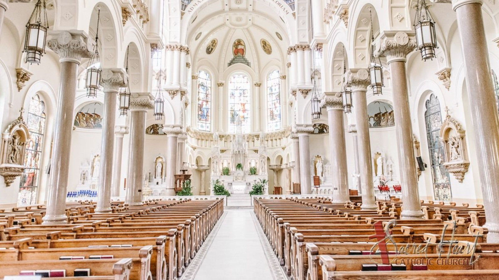 Interior of a grand church with rows of wooden pews, white pillars, and stained-glass windows.