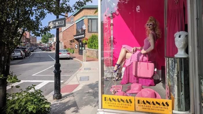 Storefront window display featuring a mannequin in pink attire, on a street corner, with town buildings visible.