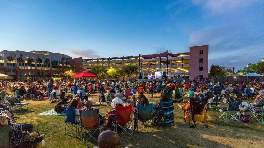Crowd seated on lawn at outdoor concert, stage in background. Buildings, blue sky, chairs, blankets.