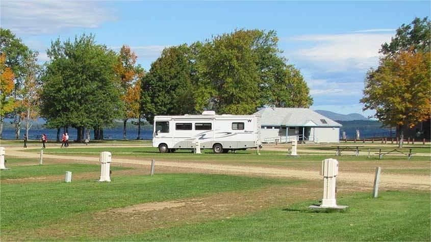 RV parked at lakeside campground with trees and building on a sunny day.