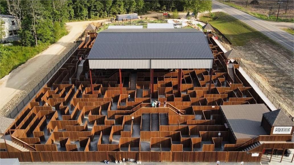 Aerial view of a large wooden maze with a covered central area, surrounded by trees and a building.
