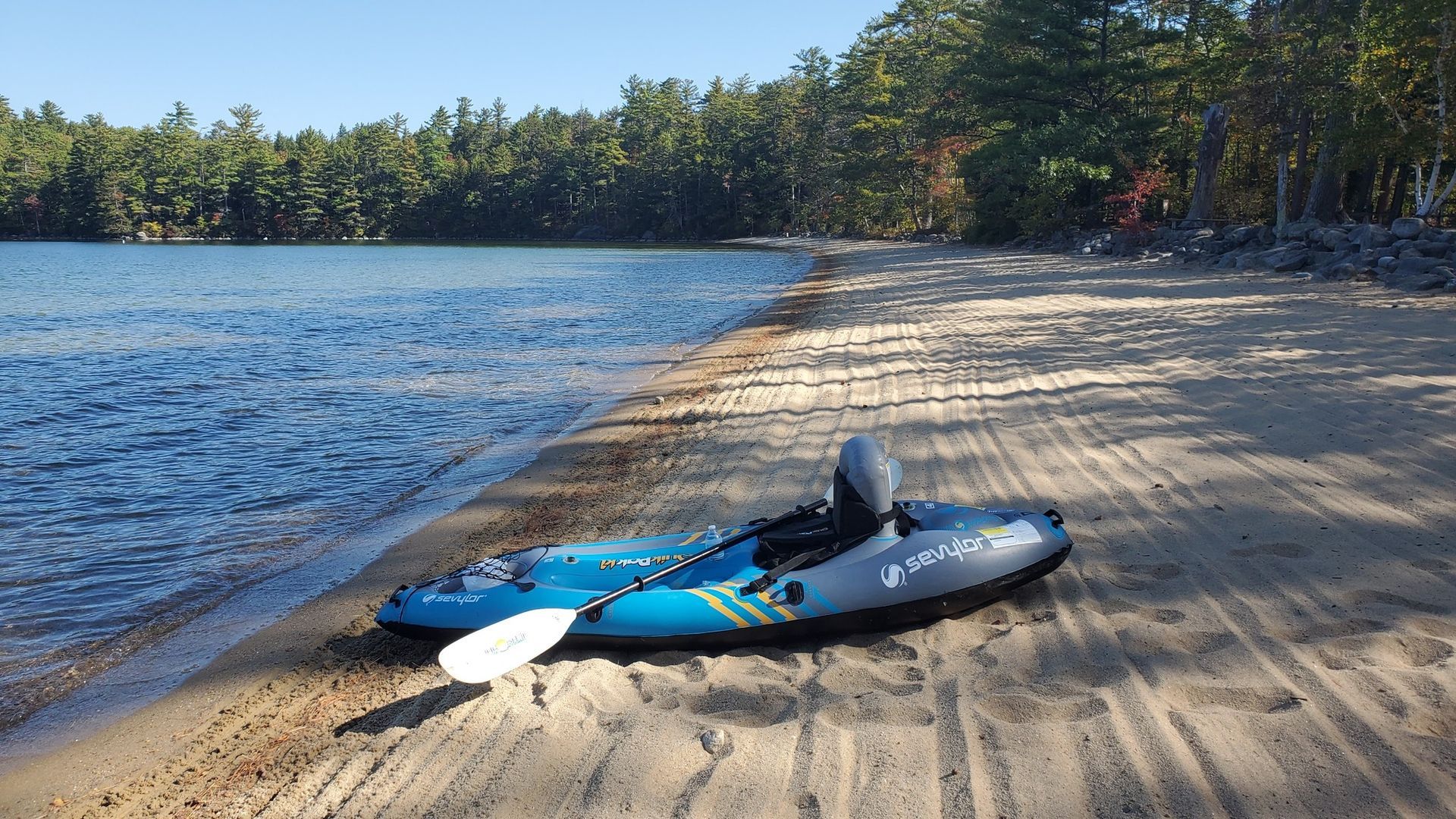 Kayak on sandy beach at lake, with paddle. Blue water, trees in the background on a sunny day.