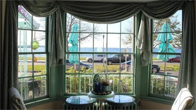 View through a bay window with green trim overlooking a street and water; draped green curtain.