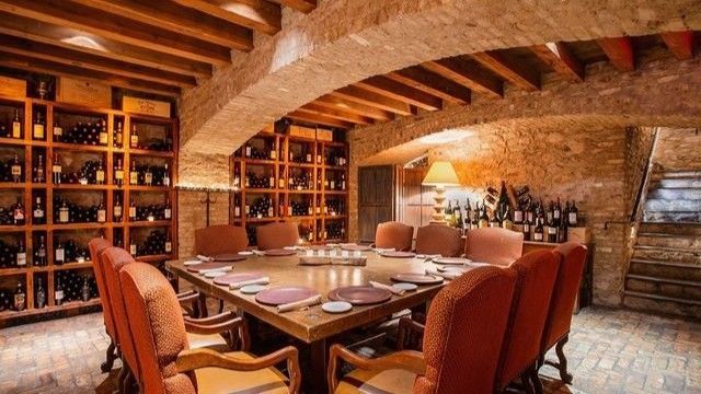 Wine cellar dining room, brick walls, wooden shelves with wine bottles, large square table with chairs.