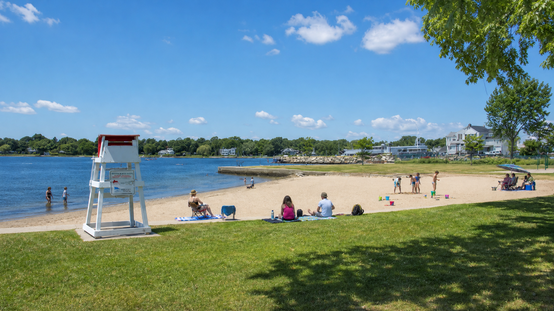 Sunny lakeside beach with people lounging on sand, blue water, and a lifeguard chair under a clear sky