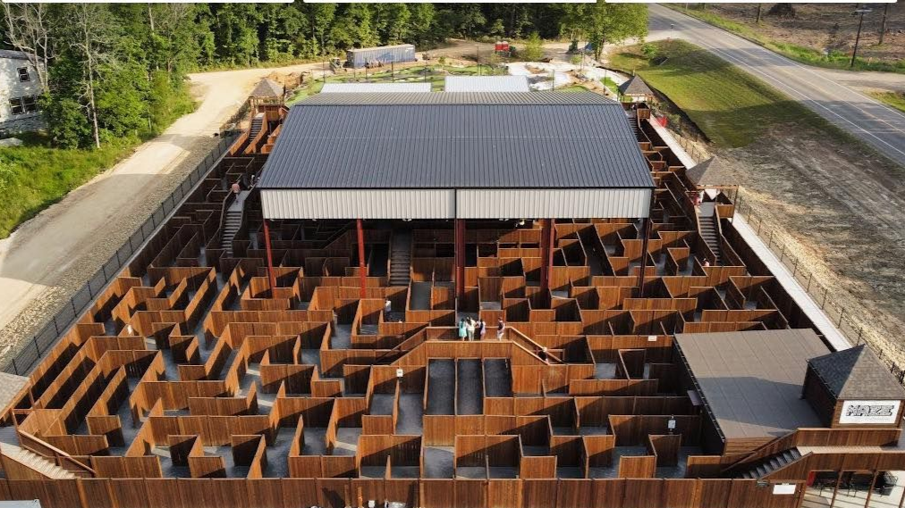 Aerial view of a large, wooden maze with a covered structure in the center; surrounding by a walkway.