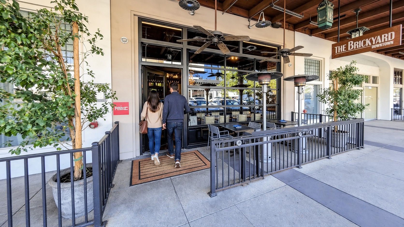 Outdoor café entrance with black railings, potted greenery, and two people walking in.