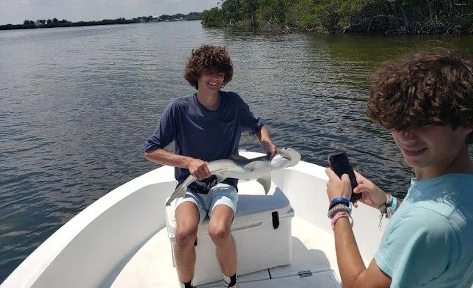 Two boys on a boat holding a fish, one taking a photo. Sunny day, water, green trees in background.
