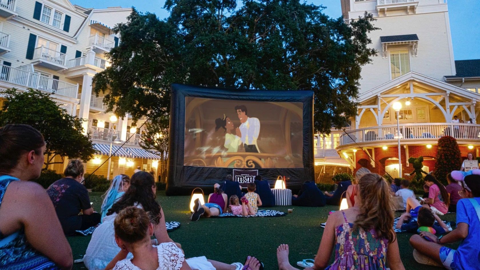 Outdoor movie screen with people watching in front of a building at dusk.