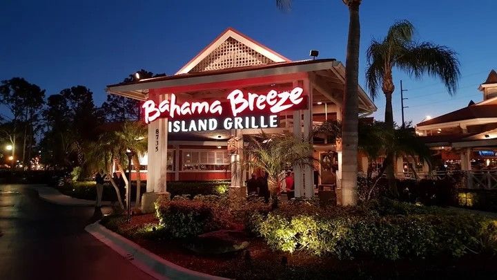 Bahama Breeze Island Grille restaurant at dusk; red illuminated sign, palm trees, and building exterior.