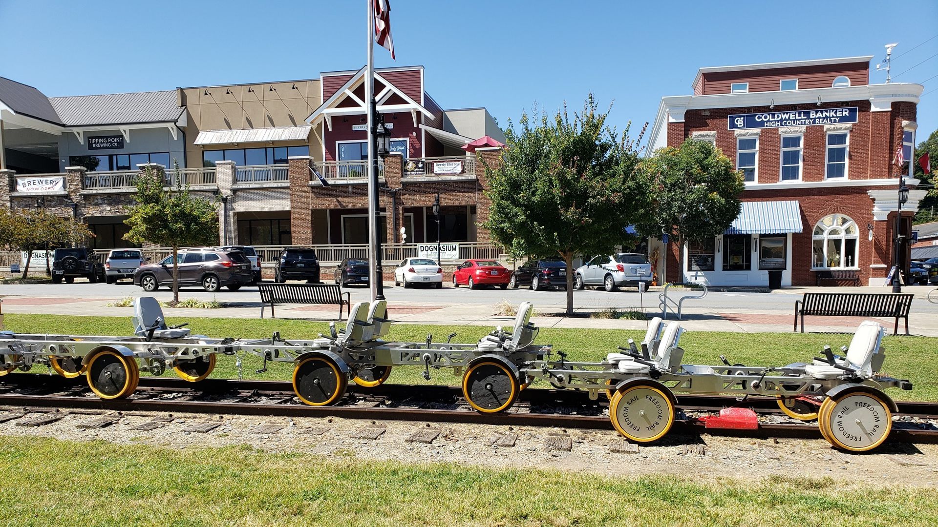 A row of rail bikes sits on tracks in front of small-town brick buildings and parking on a sunny day.
