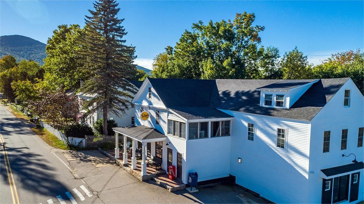 White buildings with dark roofs on a street, trees in background, mountains visible.