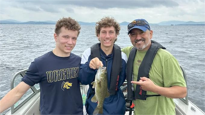 Three people on a boat, one holding a fish; two wearing life vests, posing with smiles, lake backdrop.