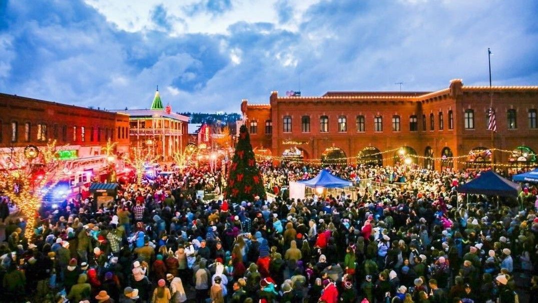 A large crowd gathers in a town square at twilight, illuminated by festive lights and a central Christmas tree.
