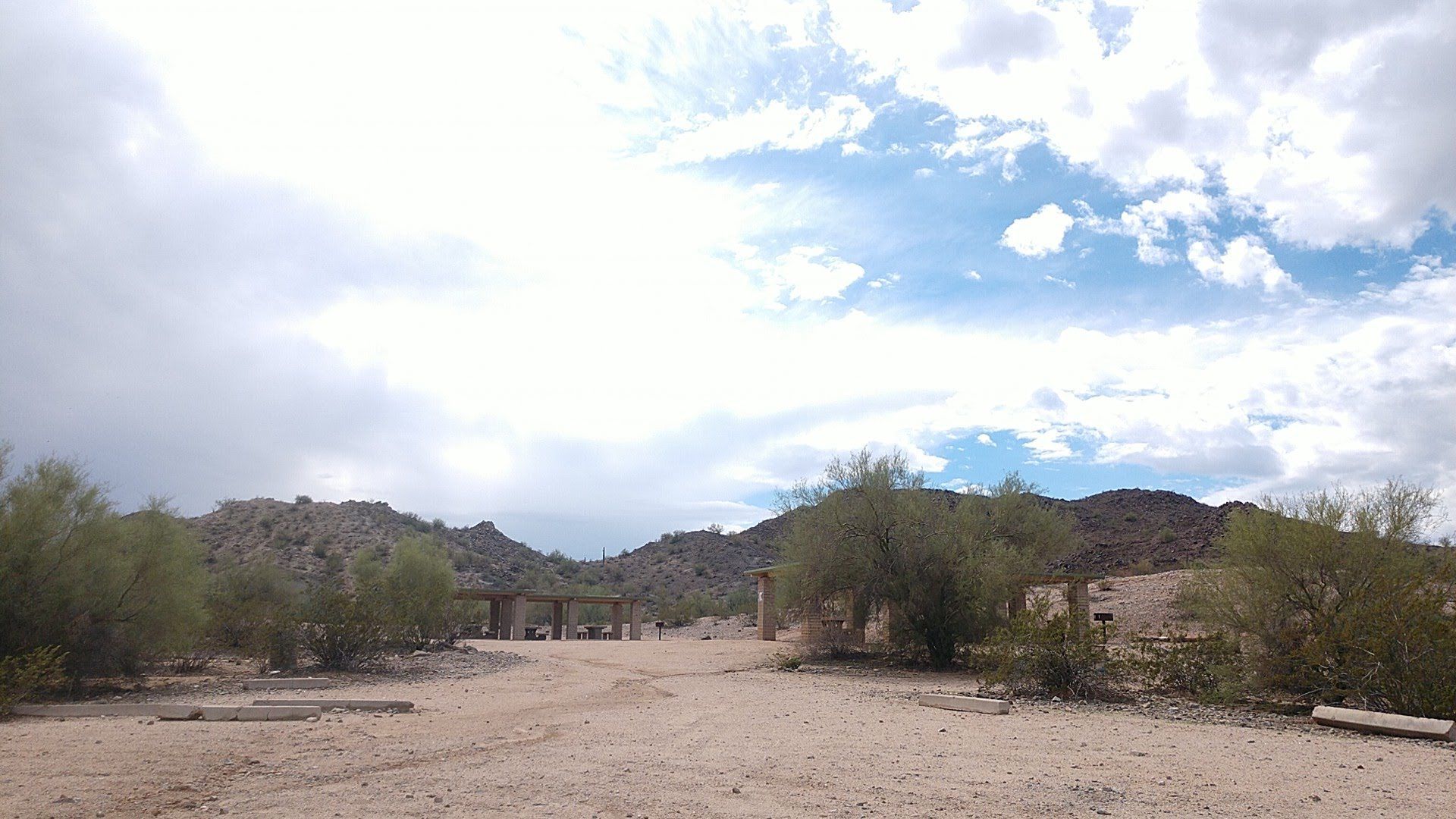 Desert landscape with cloudy sky. Dirt road, sparse green bushes, rocky hills.
