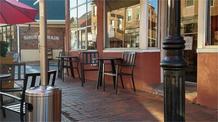 Outdoor cafe seating area next to a building with large windows. Tables, chairs, and a red umbrella are visible.