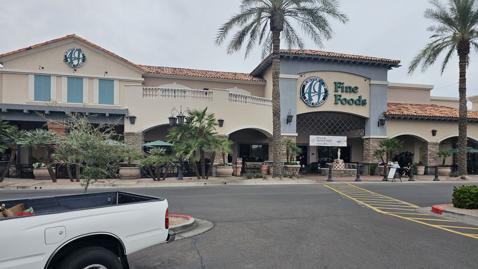 Exterior of a Fine Foods grocery store and adjacent shops, under overcast skies. A white truck is parked in the foreground.