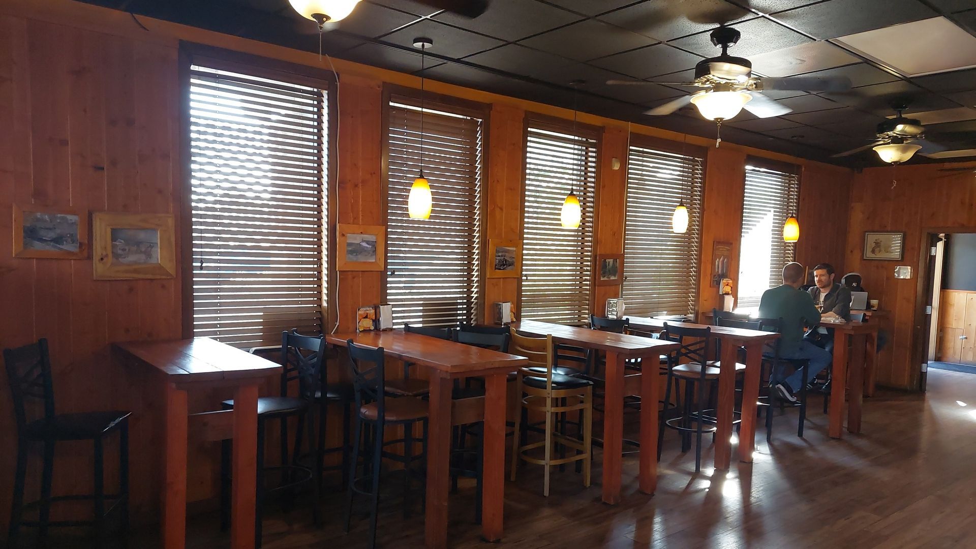 Interior of a restaurant with wooden paneling, tables, and bar stools near windows with closed blinds. People are present.