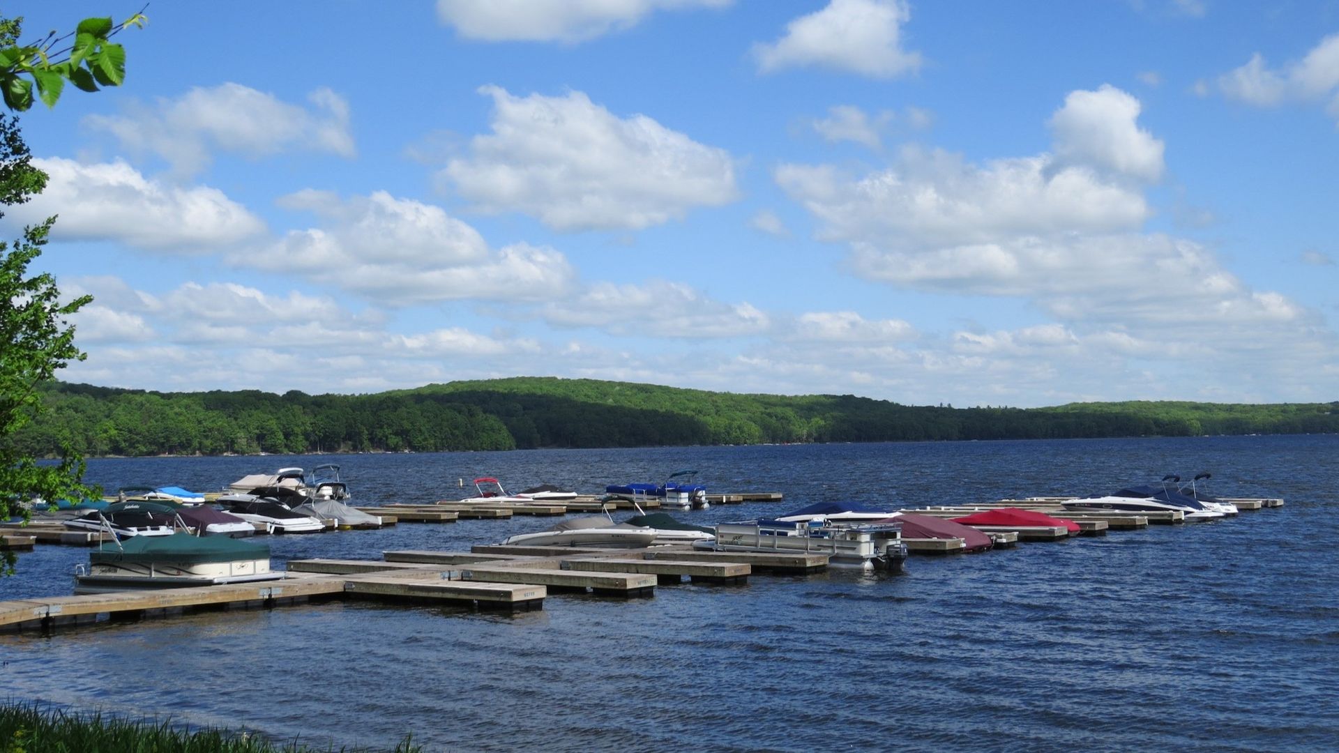 Boats docked at a lake pier with a blue sky, white clouds, and green forested hills in the background.