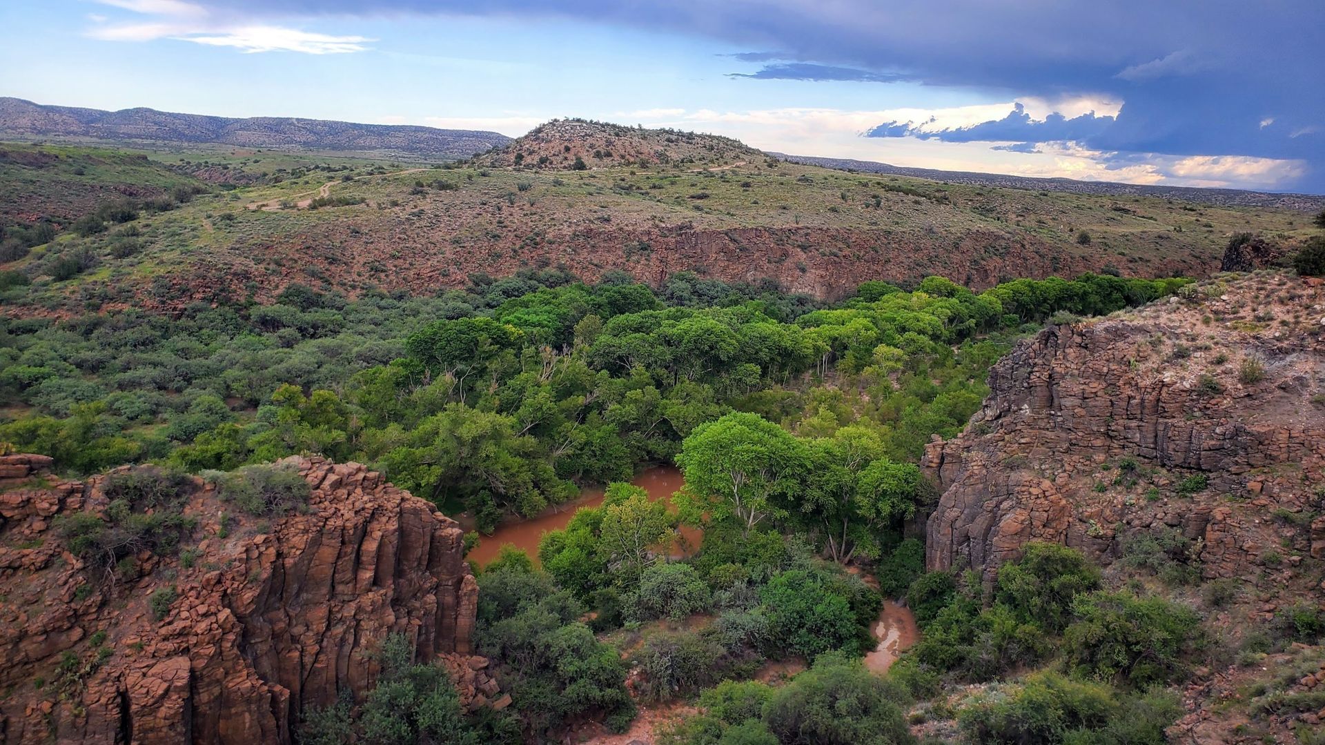 View of a canyon with red rock walls and lush green vegetation in the center; hills and cloudy sky in the background.