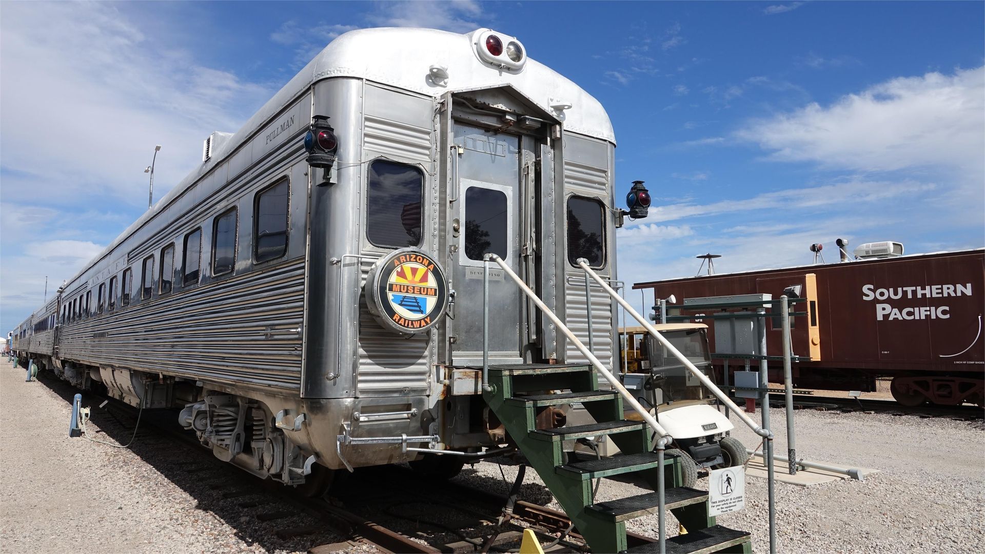 Silver train car with steps, Arizona state seal, sunny sky, and Southern Pacific freight car.