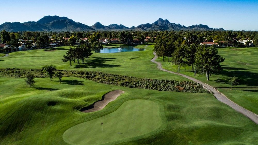 Green golf course with a small pond, trees, and mountain backdrop.