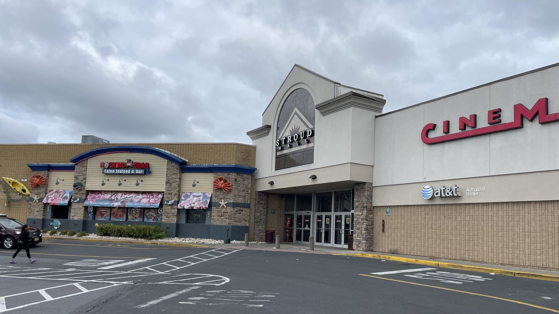 Exterior of a shopping center entrance and a cinema building on a cloudy day with a parking lot in the foreground.