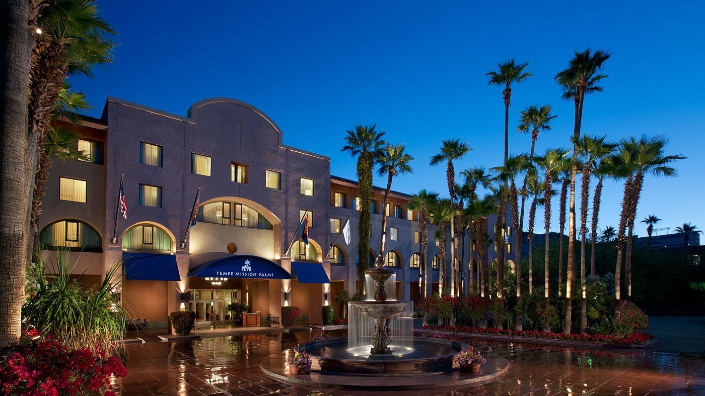 Hotel entrance at dusk, fountain, palm trees, blue awning, and building with lights.