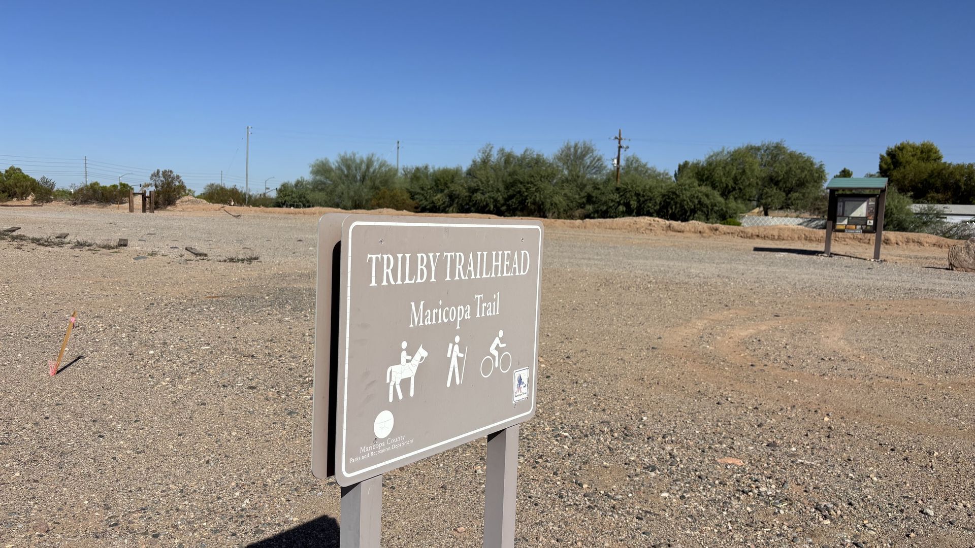 Trailhead sign in a gravel area under a blue sky, with icons for horses, walkers, and cyclists.