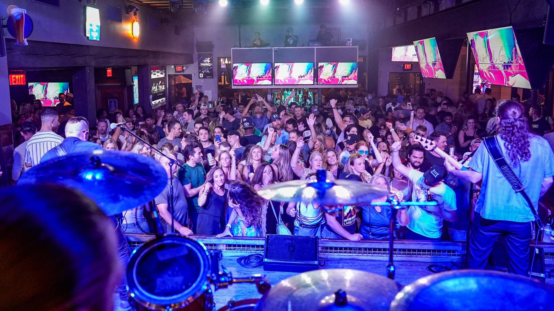 Band playing on stage in a crowded bar. Audience with raised hands, bathed in blue stage light.